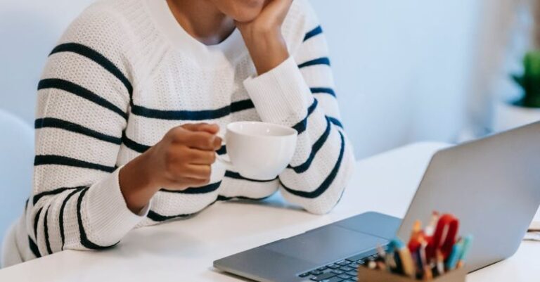 Mobile Content - Positive young African American female freelancer in casual clothes talking on smartphone and drinking coffee while working remotely on laptop at home