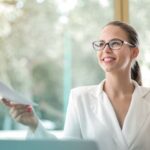 Content Success - Low angle of successful female executive manager in classy style sitting at table with laptop in contemporary workplace and passing documents to colleague