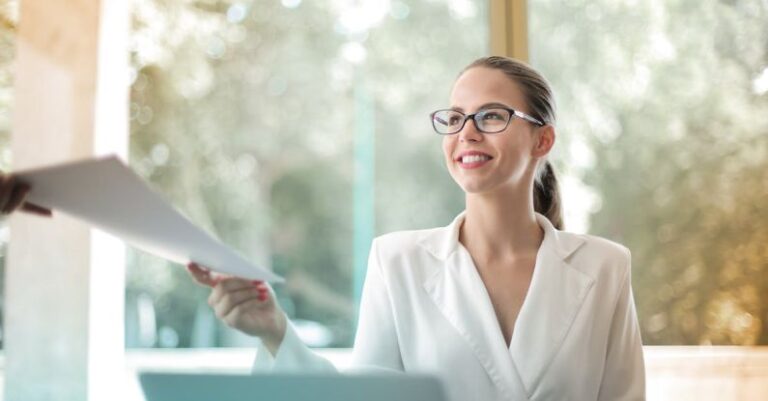 Content Success - Low angle of successful female executive manager in classy style sitting at table with laptop in contemporary workplace and passing documents to colleague