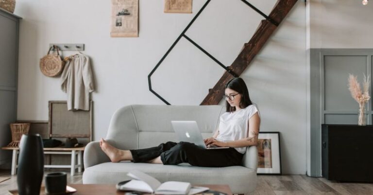 Email Content - Positive female with tattooed arms wearing casual clothes and eyeglasses reading netbook while sitting barefoot on comfortable sofa behind table with books and magazines in modern apartment with minimalist interior in eco style during free time